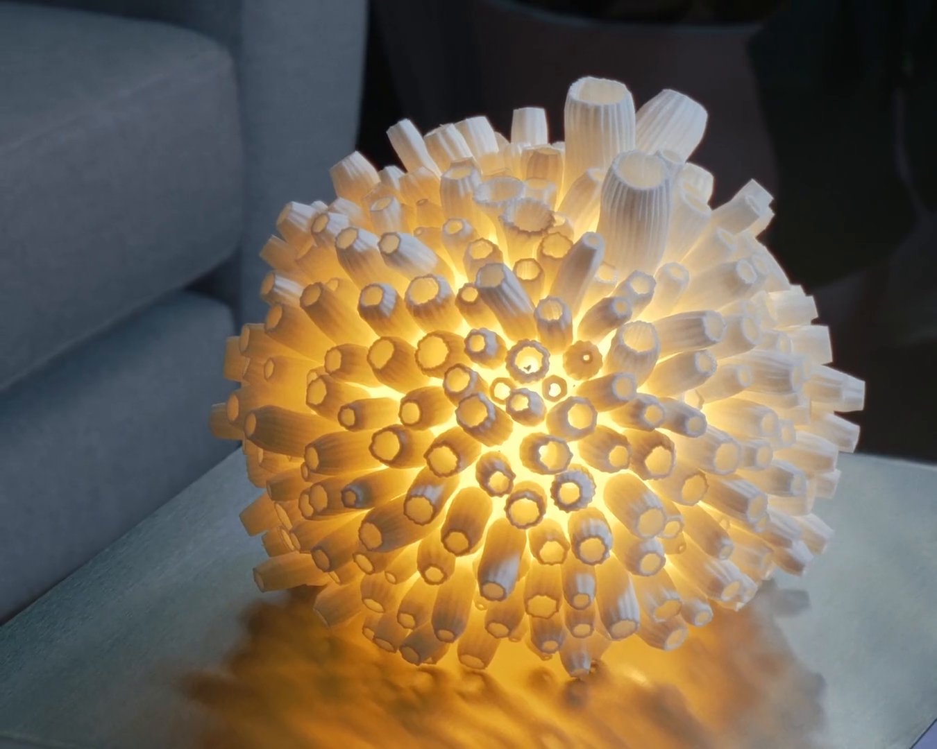 A 3D printed Bloom lamp with the light on, glowing warm through hundreds of white petals, on a table beside a sofa.